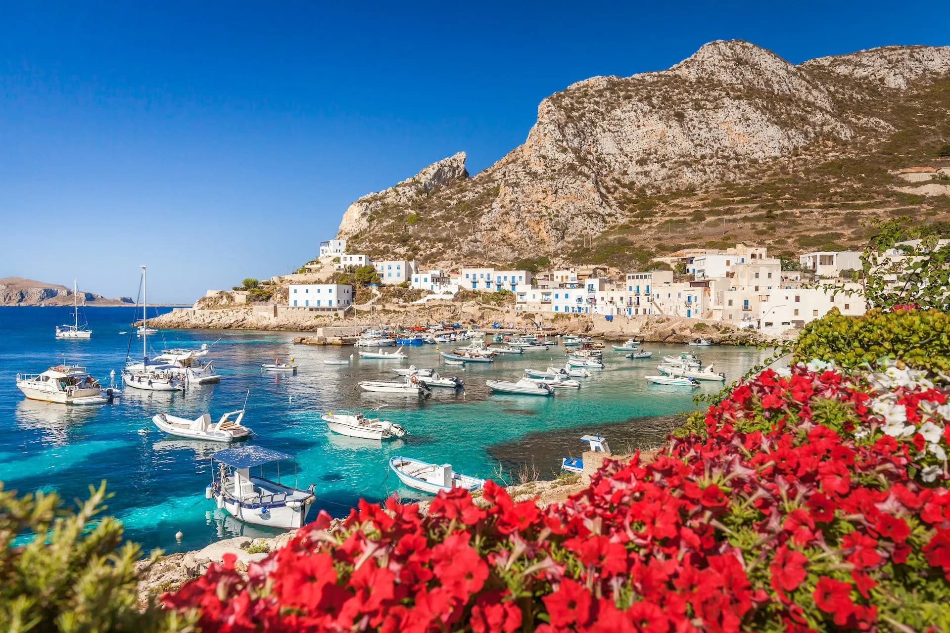 Boats anchored in turquoise harbor below white buildings and arid mountain, Levanzo.