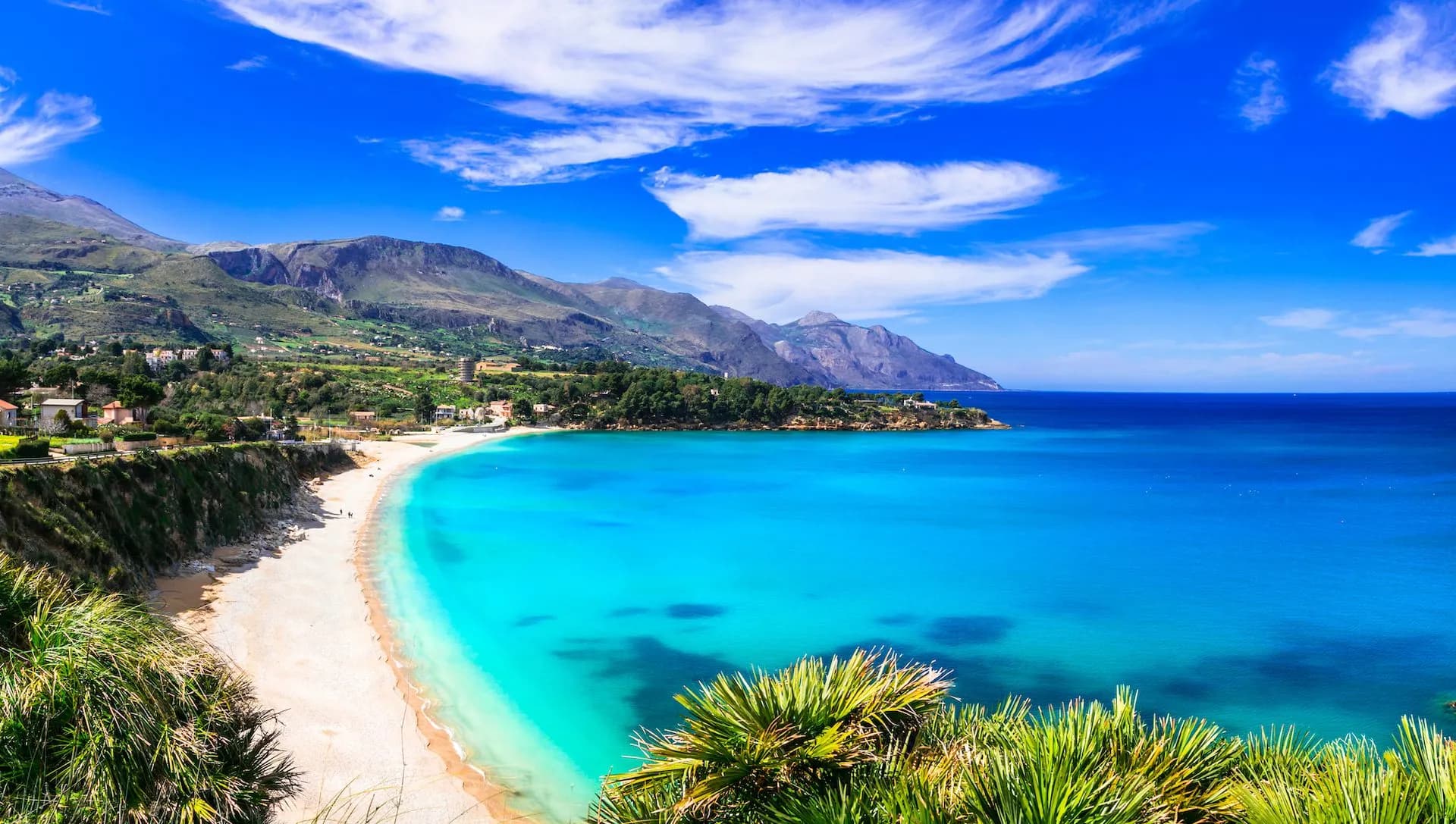 Sandy beach curving into turquoise Mediterranean Sea with mountains in Scopello