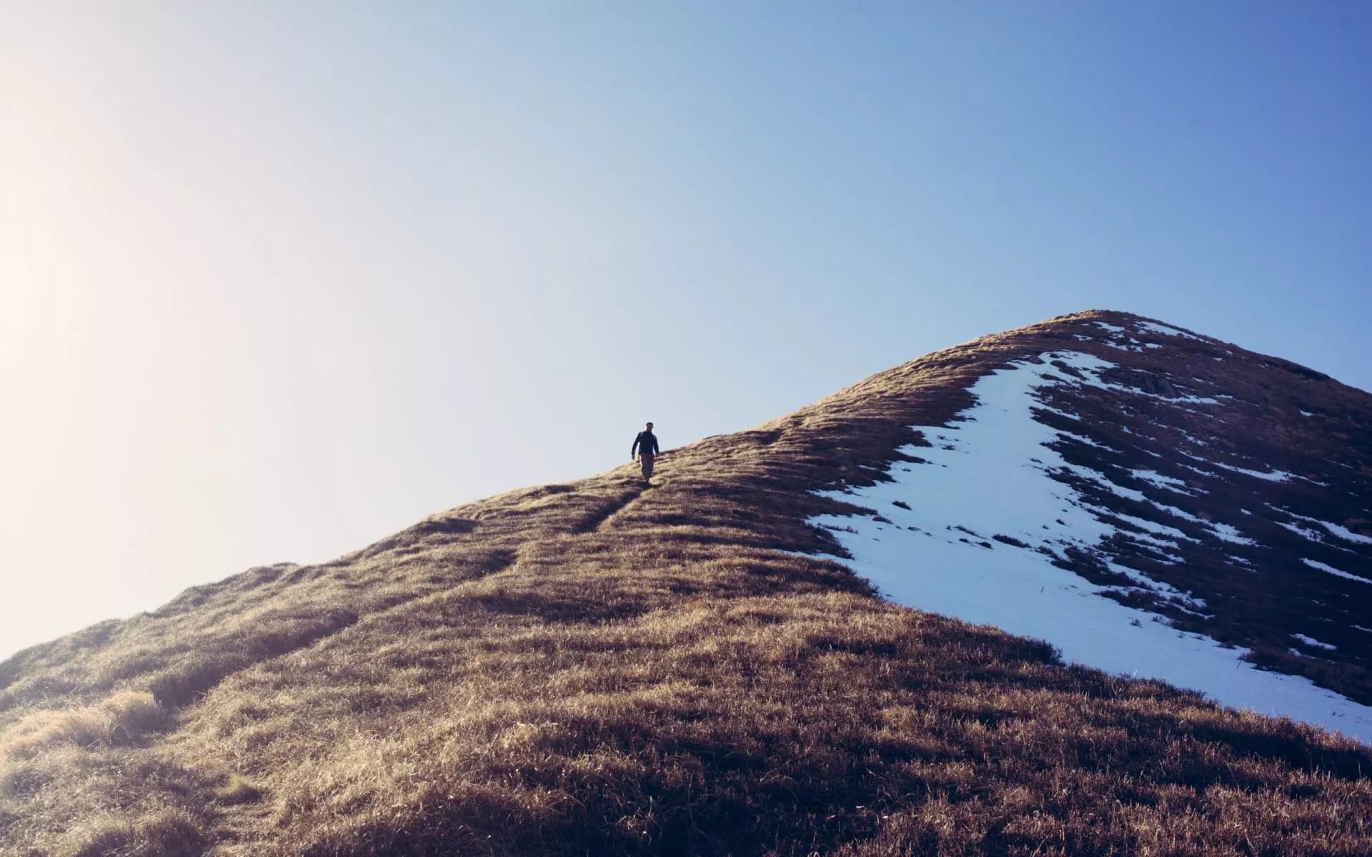 Hiker ascending a grassy, partially snow-covered mountain ridge under a clear blue sky.