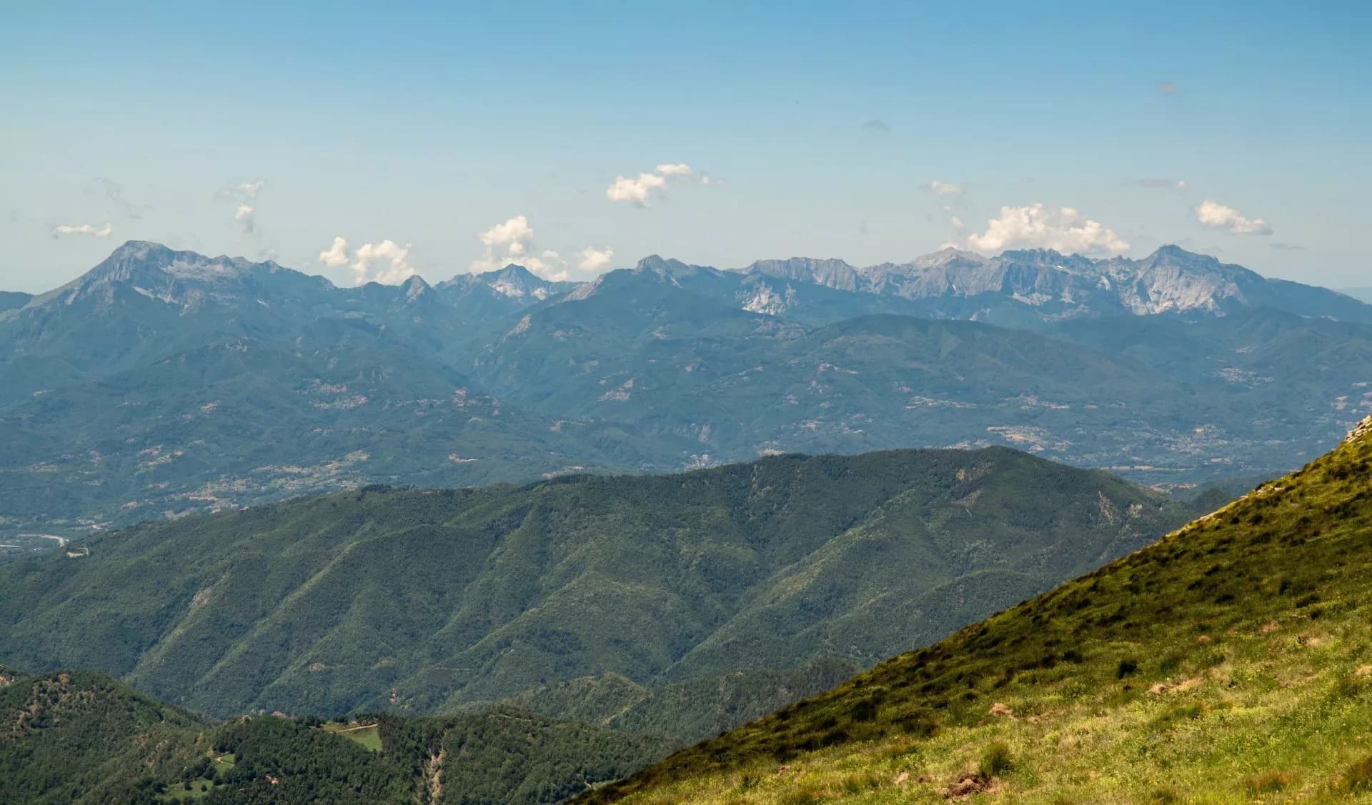Mountain range panorama with green slopes in foreground under blue sky in Alpi Apuane National Park.