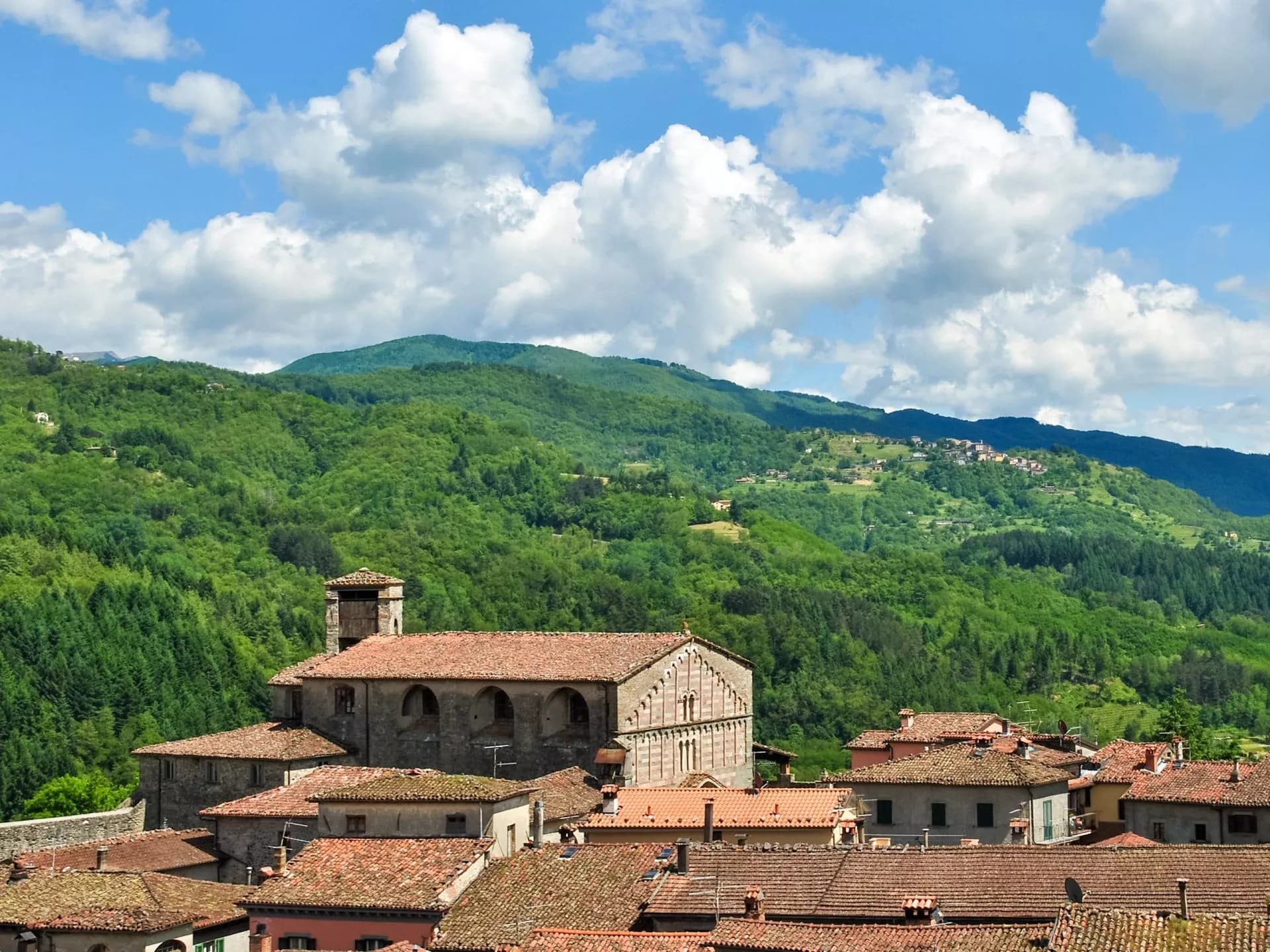 Roofs of Castiglione di Garfagnana town with stone church and lush green mountains under blue sky.