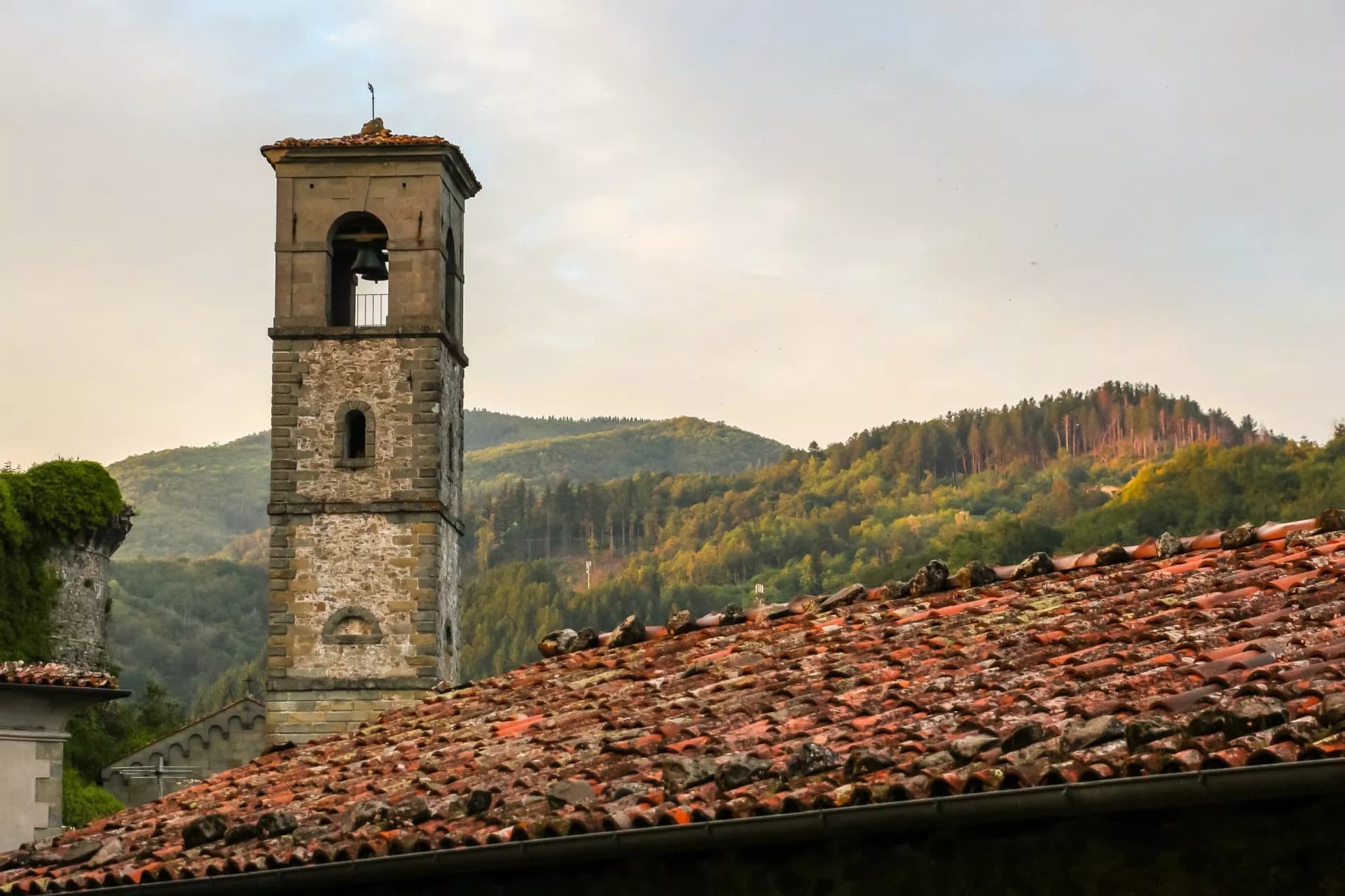 Stone bell tower above terracotta roofs with forested hills in Castiglione di Garfagnana.