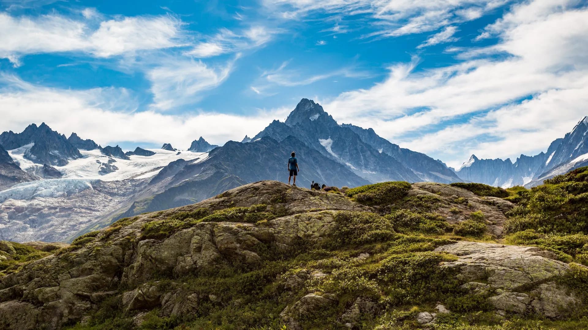 Hiker standing on rocky outcrop overlooking massive snow-capped mountain range under blue sky