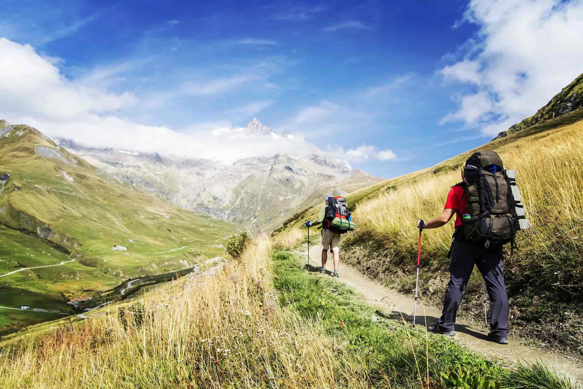 Hikers with large backpacks trekking on a dirt path toward snow-capped mountains under blue sky.