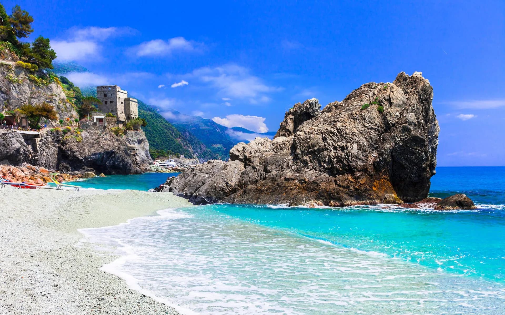 Pebble beach with turquoise water, large rock formation, and historic tower in Monterosso, Italy.