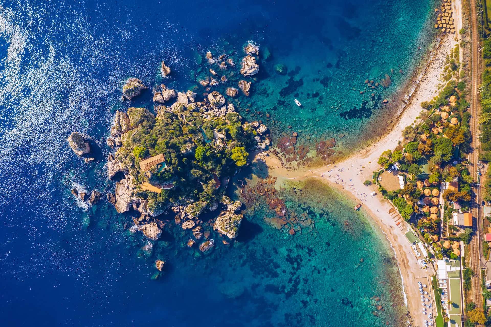 Aerial view of Isola Bella island connected to a beach with turquoise water and nearby railway line in Sicily.