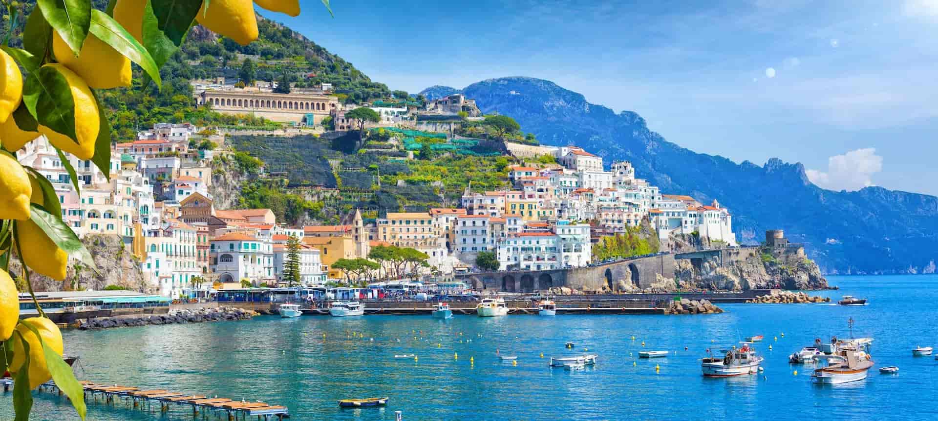 Amalfi town on cliffside with colorful buildings, boats in blue water, and lemons in foreground.