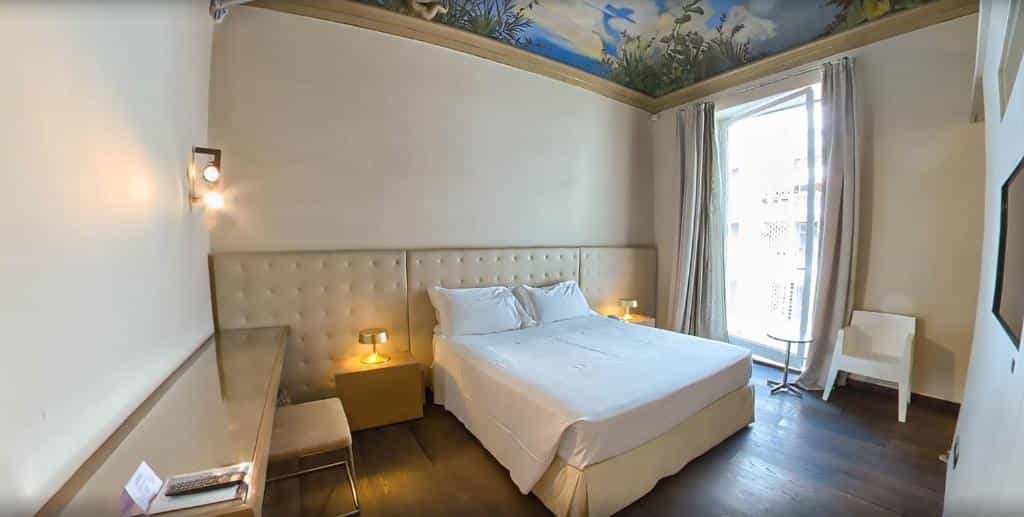 Hotel Romano House bedroom with white bedding, mural ceiling, and bright window view.