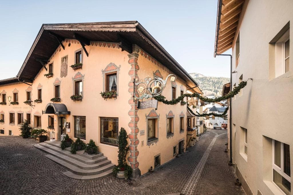 Traditional hotel building with painted facade and festive garlands on a cobblestone street in an Alpine village.