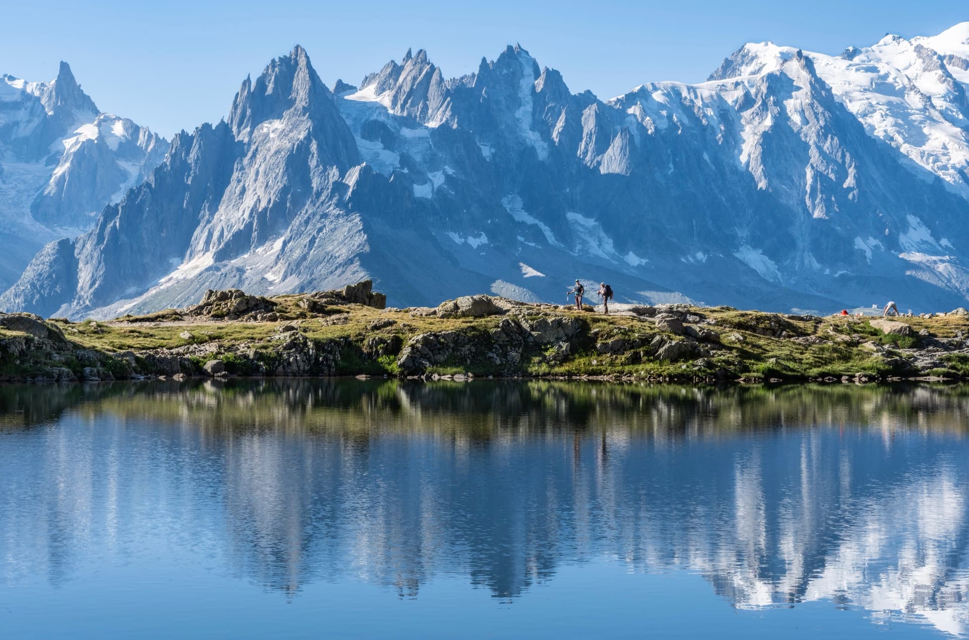 Hikers by alpine lake reflecting jagged snow-covered mountains