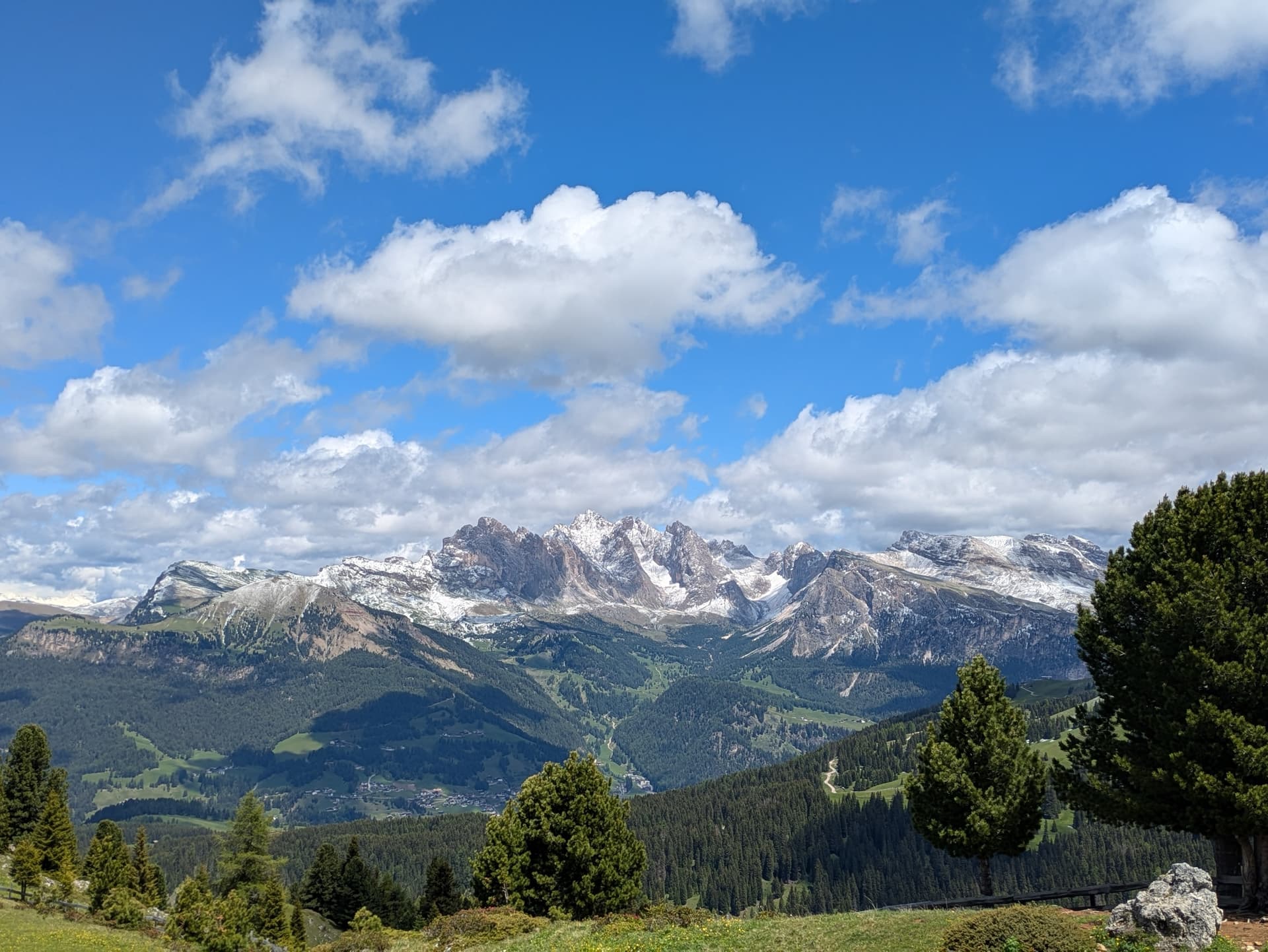 Snowy mountain range across forested hills under a blue sky with white clouds