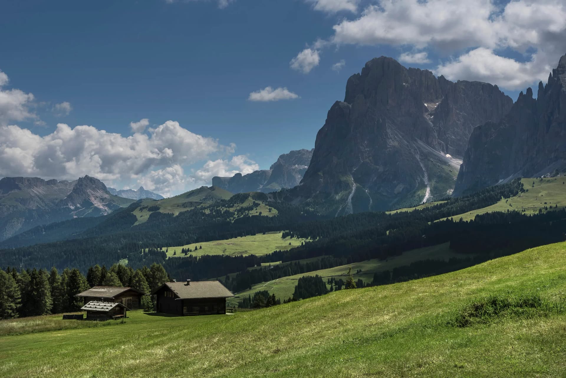 Alpine huts on green meadow with massive rocky mountains under blue sky and clouds.